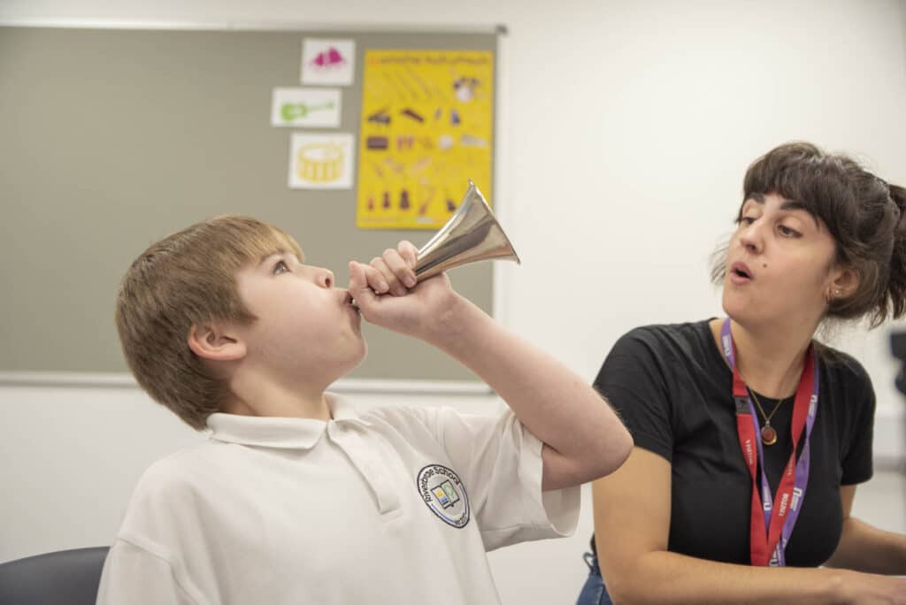 Charlie looks to the ceiling as he blows into a small silver horn, with music therapist Stella singing alongside him.