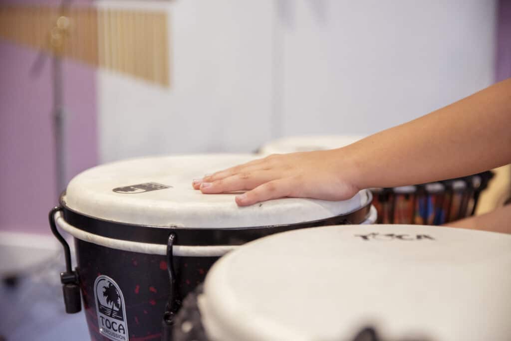 A person reaches out their hand to play music on a drum, which sits in a room full of musical instruments