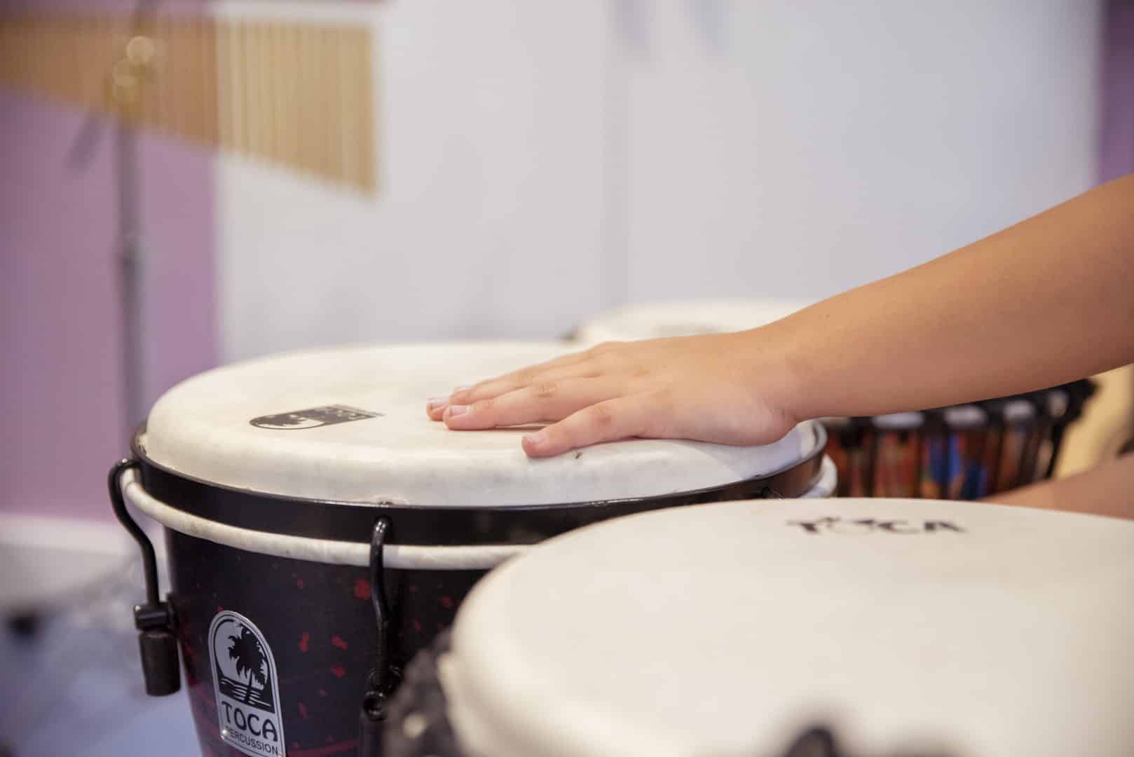 A person reaches out their hand to play music on a drum, which sits in a room full of musical instruments