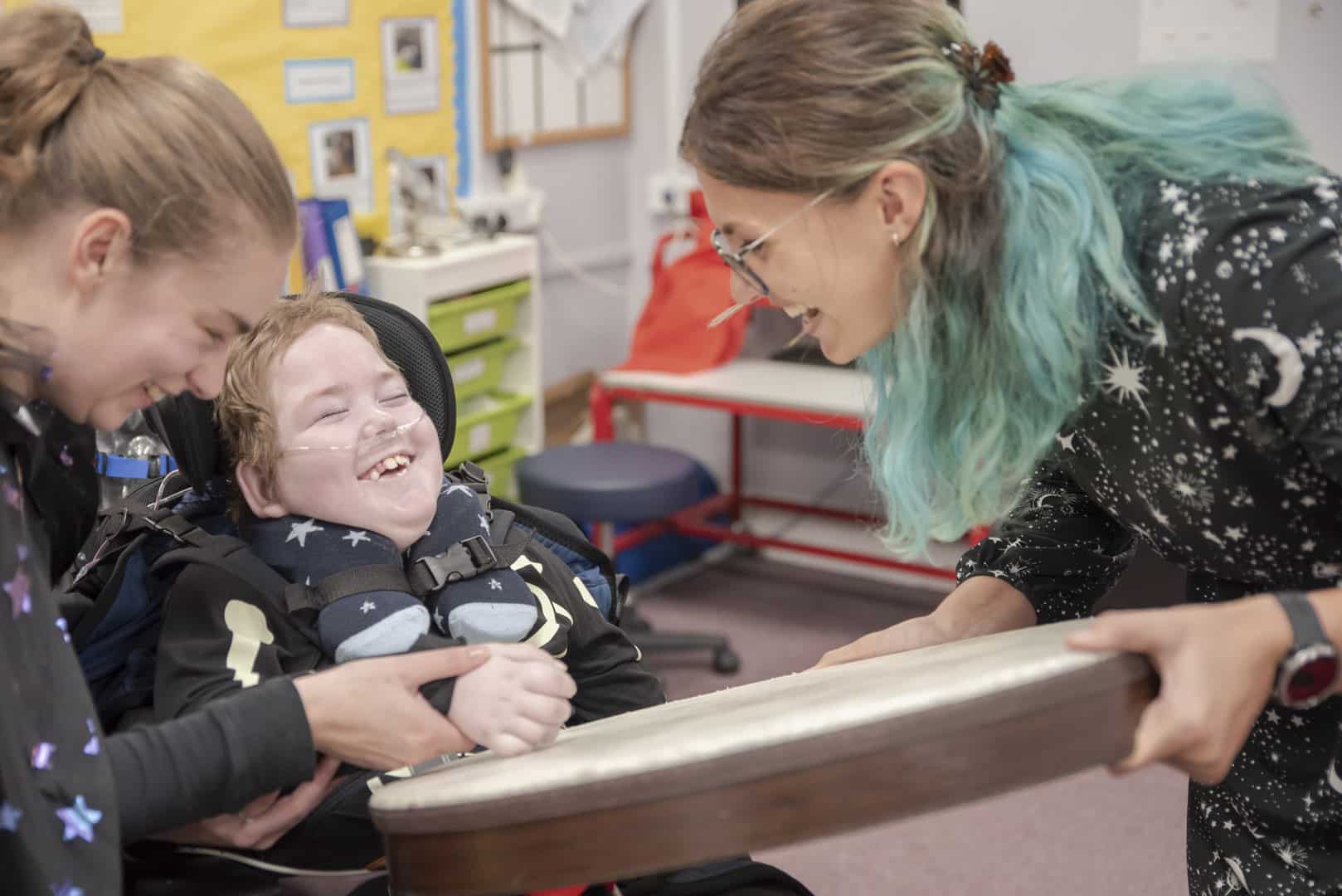Two people hold out a drum to a young client during music therapy at a Nordoff and Robbins partner school. Together, they all smile and laugh.