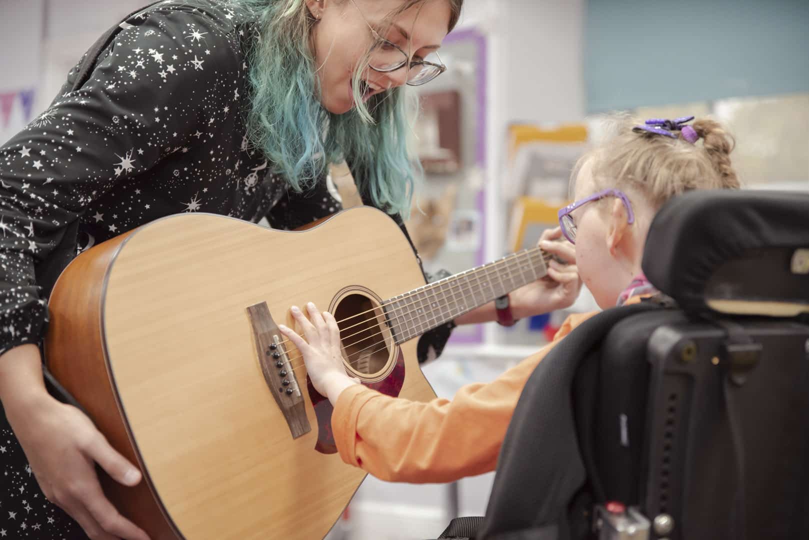 During a music therapy session, someone excitedly stands with an acoustic guitar, as a young client reaches out to play the instrument.