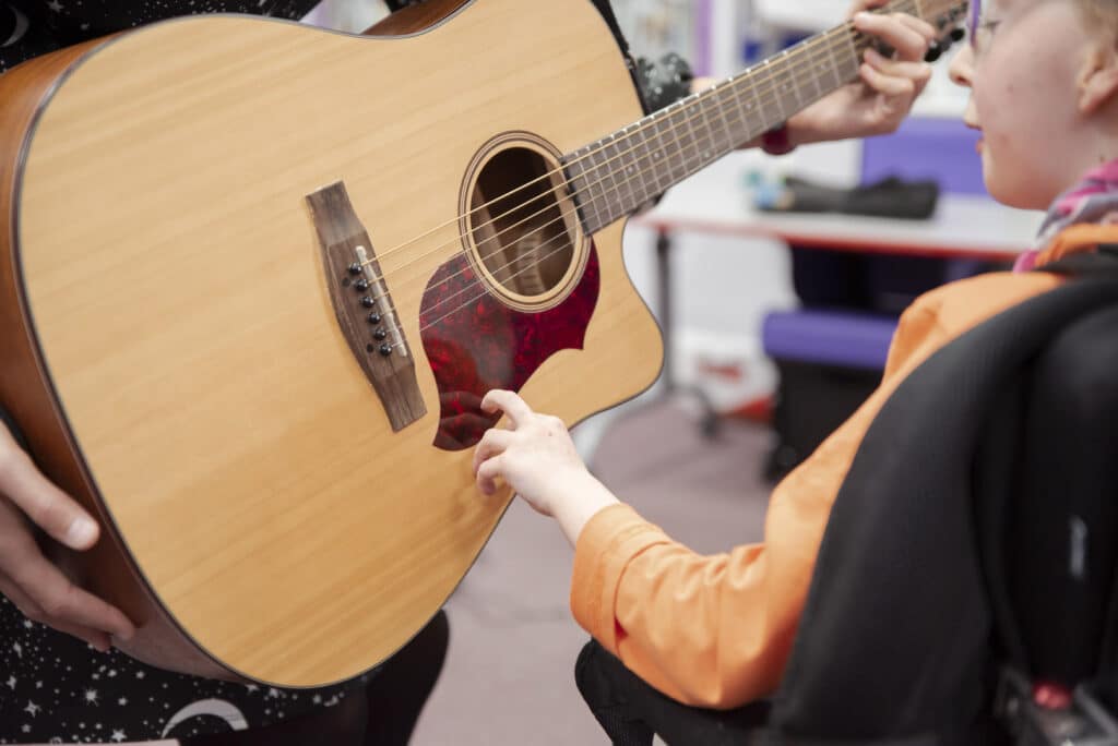 A young client reaches out towards an acoustic guitar, as someone stands up beside them to play the instrument