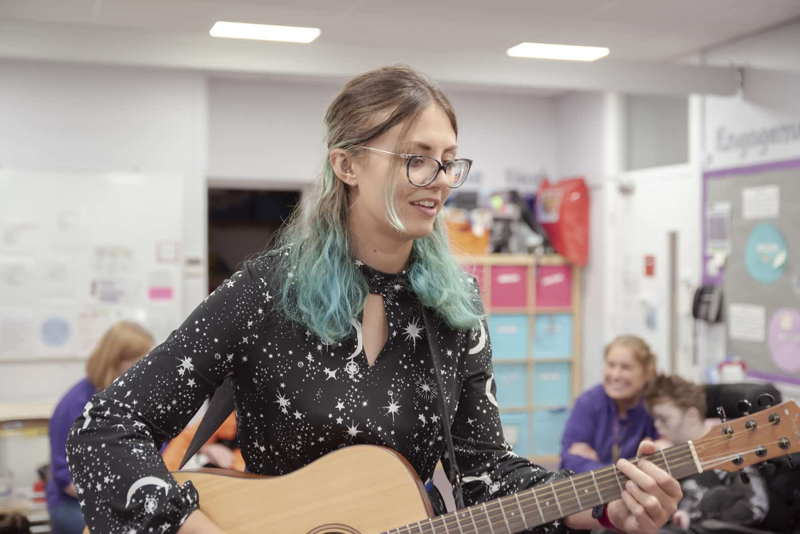A person stands to play the acoustic guitar at one of our partner organisations, as others play music around them