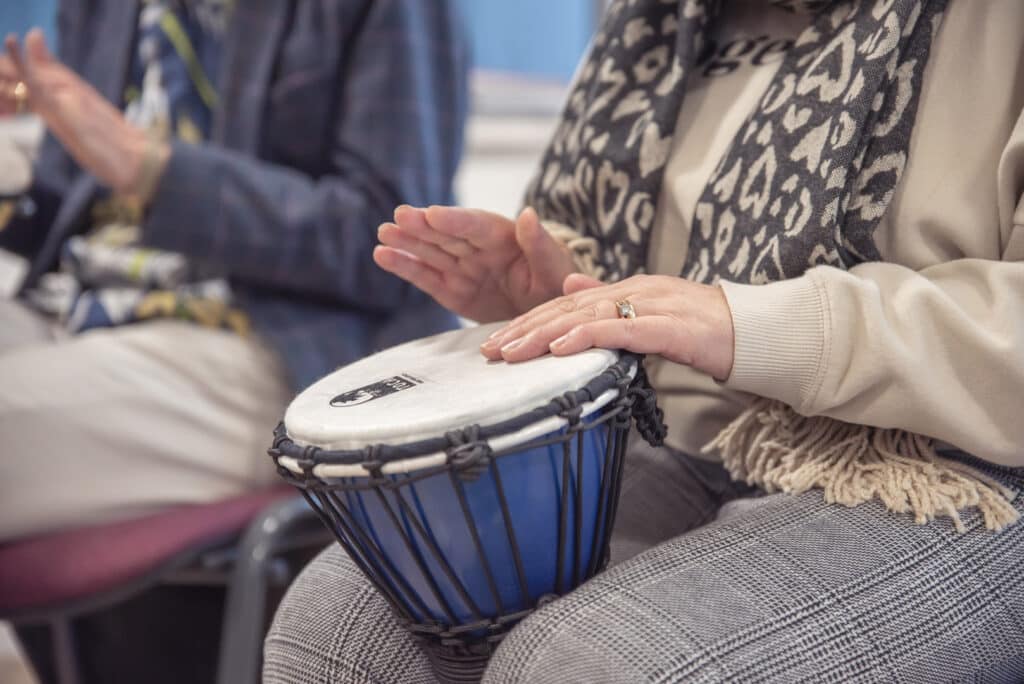 Someone sits down with other people to play music on a small drum, as they take part in a music therapy session