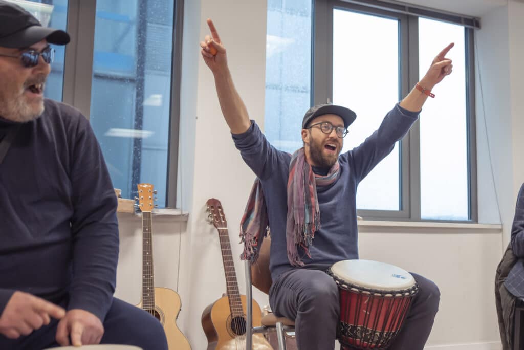 Music therapist Paddy enthusiastically points to the ceiling as he sits behind a drum. He sits amongst clients at a music therapy session at ReCoCo, one of our partner organisations.