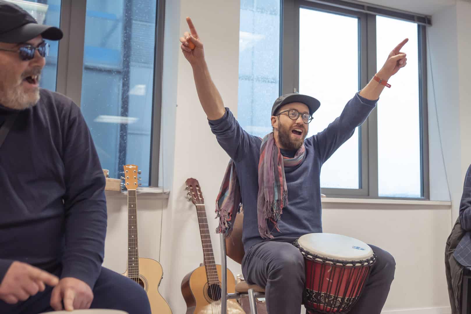 Music therapist Paddy enthusiastically points to the ceiling as he sits behind a drum. He sits amongst clients at a music therapy session at ReCoCo, one of our partner organisations.