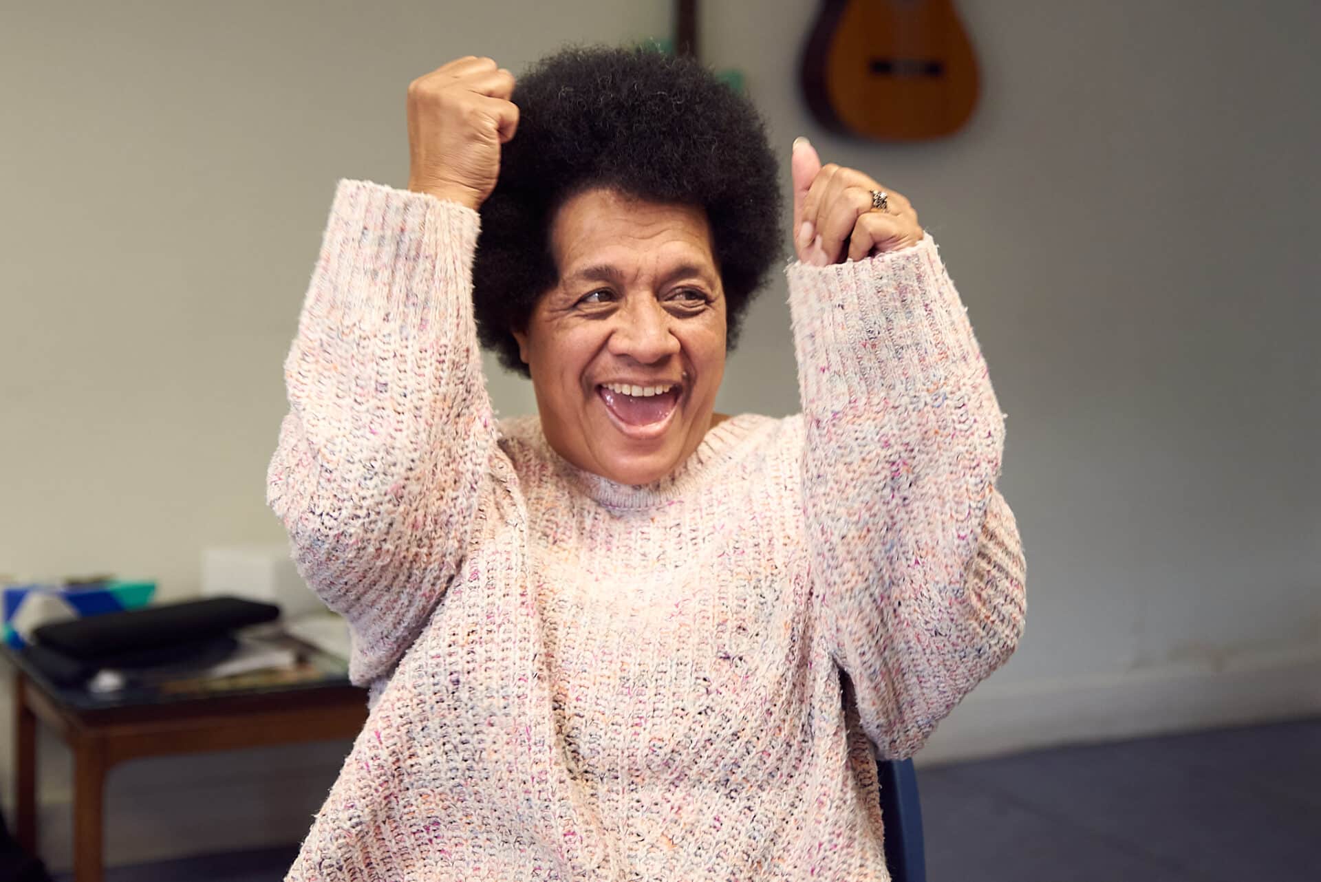 A person in a knitted jumper excitedly raises up their arms with closed fists during a music therapy session.