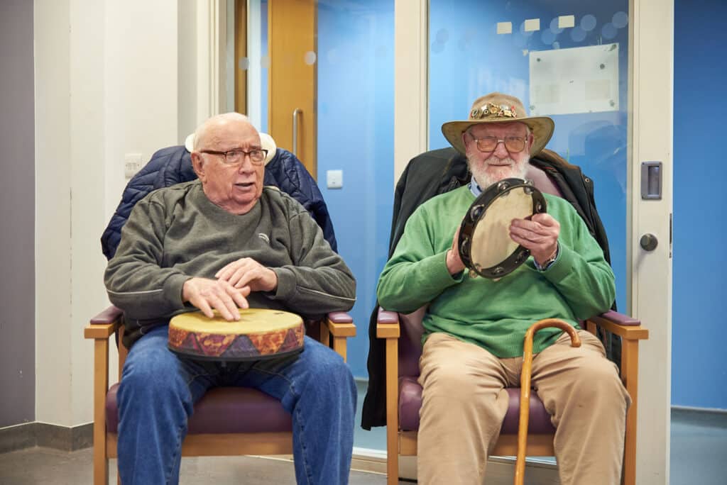 Two people sit side by side during music therapy at Dagenham Memory Clinic, making music with a drum and tambourine.