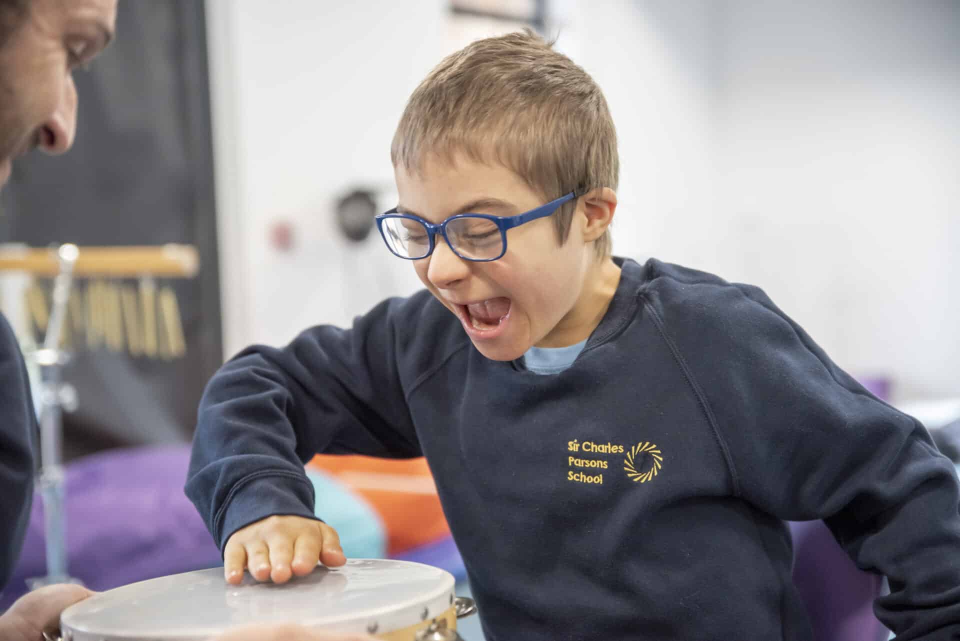 A young client excitedly creates music on a drum, as a music therapist holds the instrument out to them