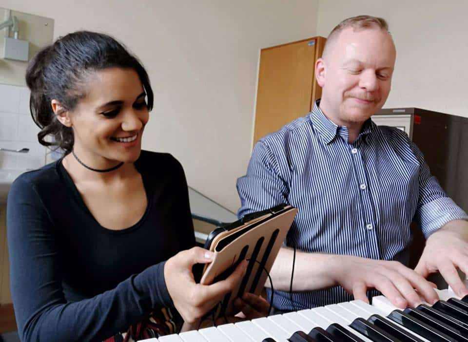 Charlotte, a Nordoff and Robbins music therapy client, smiles at an iPad next to music therapist Alan as he plays the keyboard.