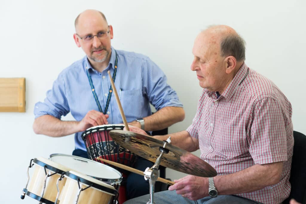 Geoff sits at a drum kit during music therapy with Fraser, his music therapist, who plays along beside him.