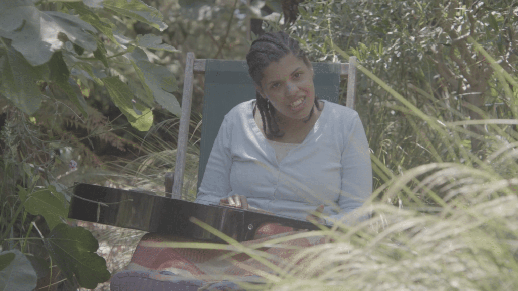 Music therapy client Inez smiles as she sits outside in the long grass, one hand on an acoustic guitar