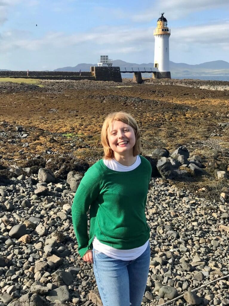 Our Supporter Care Officer Mariola smiles happily, as she stands in front of a lighthouse at the beach.