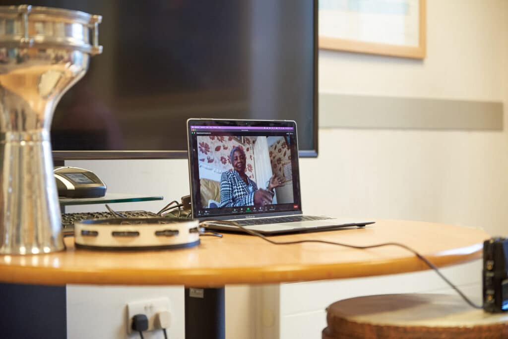An open laptop sits on a table next to tambourine. On screen, someone waves their hands as they take part in an online music therapy session.