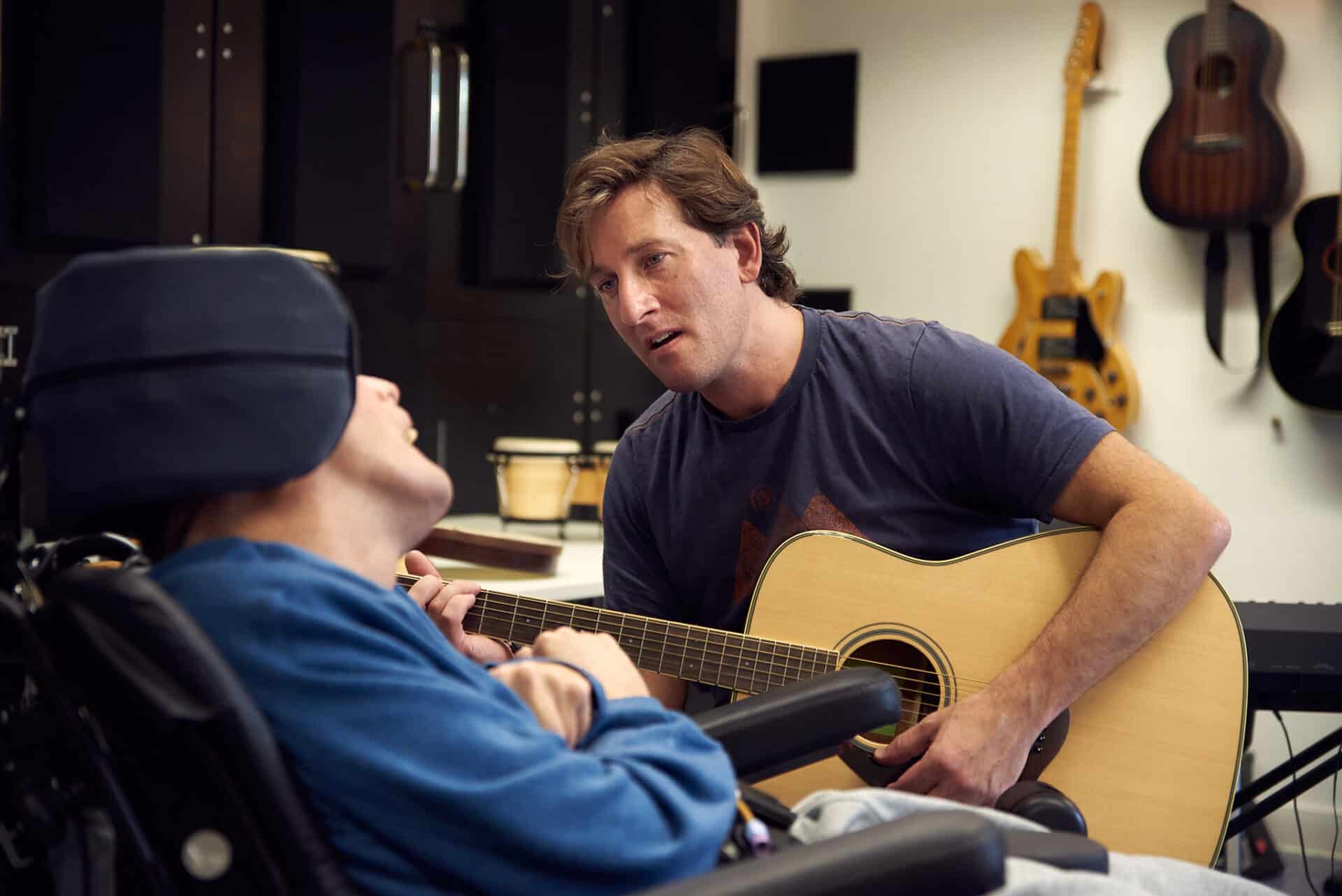 During a music therapy session, a person faces towards music therapist Oli, who is playing the acoustic guitar and singing.