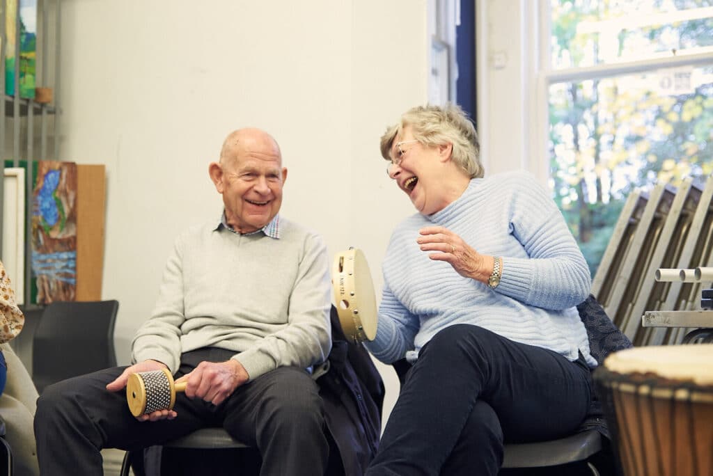 Two people laugh along together during music therapy, as they sit amongst a group playing musical instruments