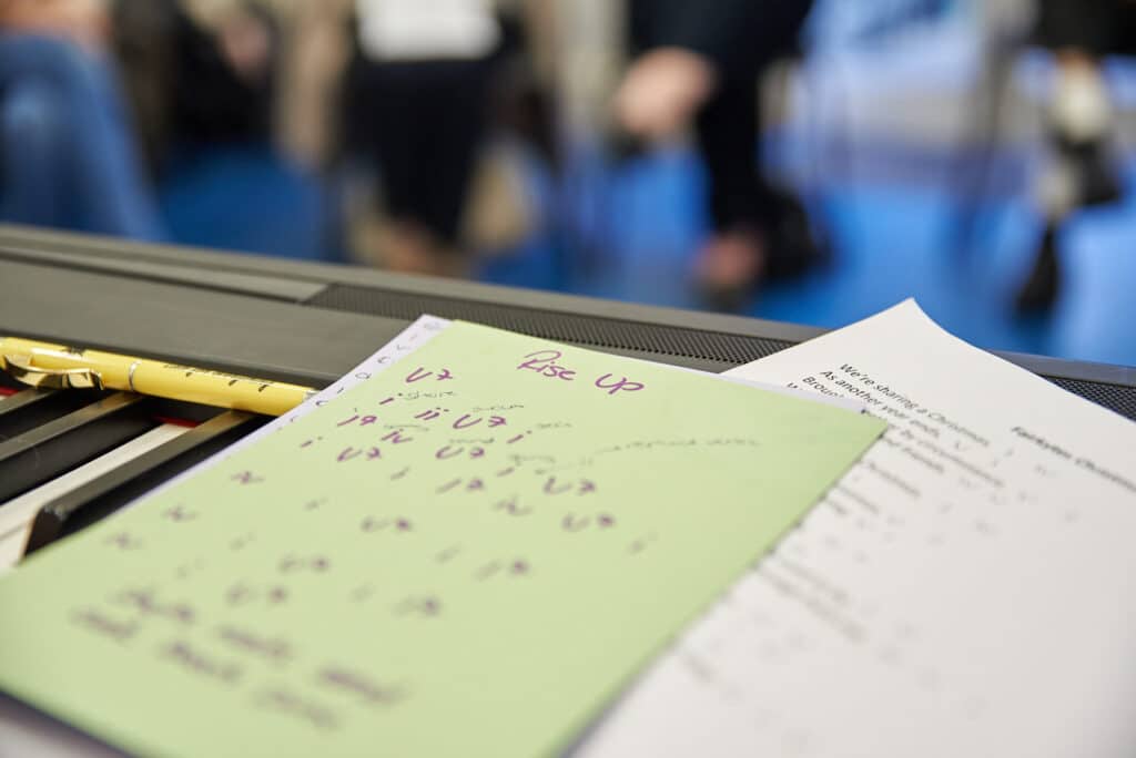 Pieces of paper sit on top of a keyboard. The first contains musical notes, with "Rise Up" written at the top. Underneath is a lyric sheet, with the first line reading "We're sharing a Christmas".