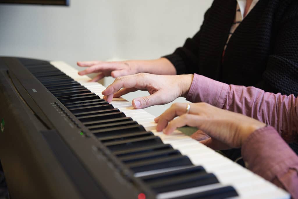 Two people place their hands on a keyboard, playing music together during a music therapy session