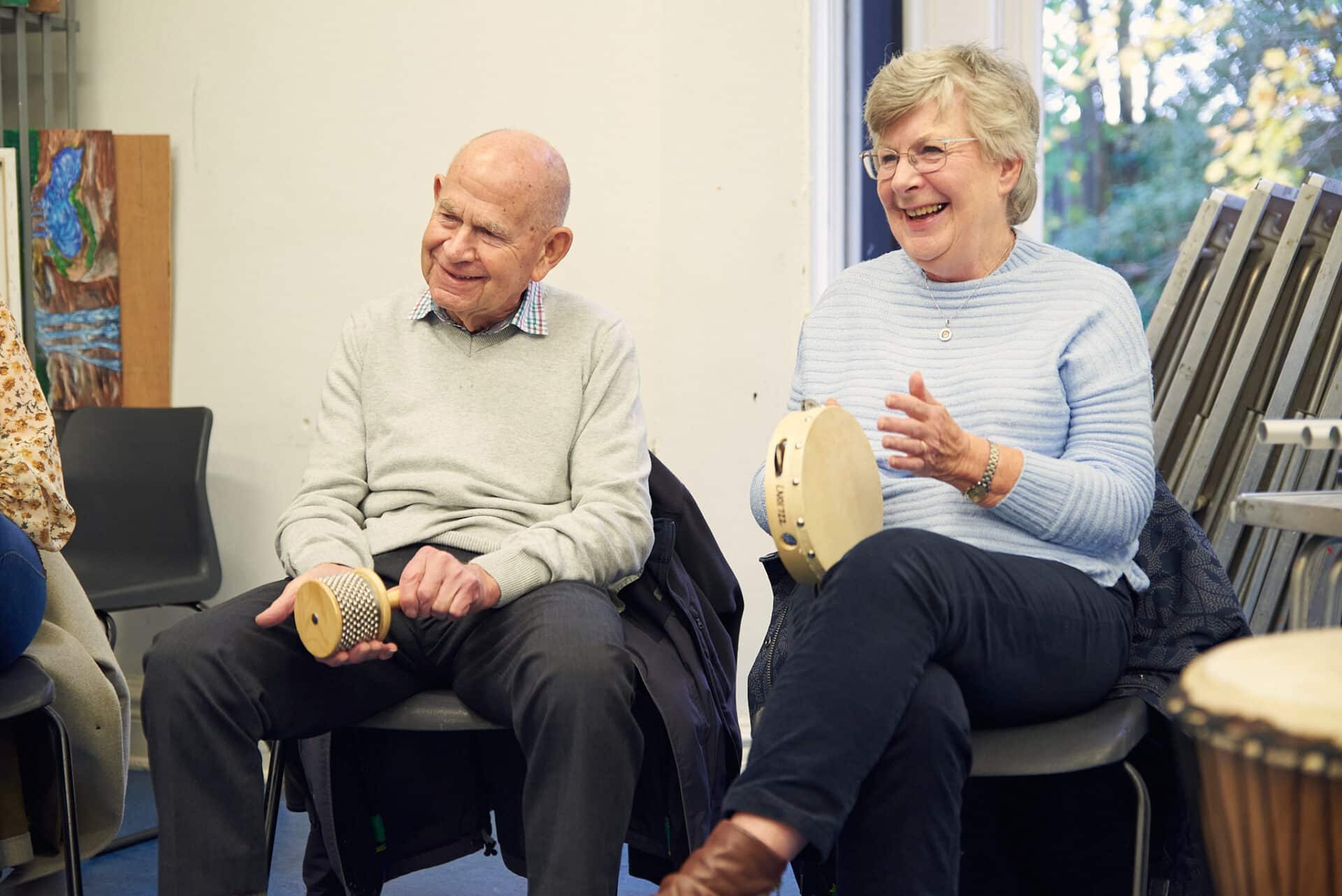 Sitting side by side, two smiling people hold musical instruments during a music therapy session at Fairkytes, one of our partner organisations.