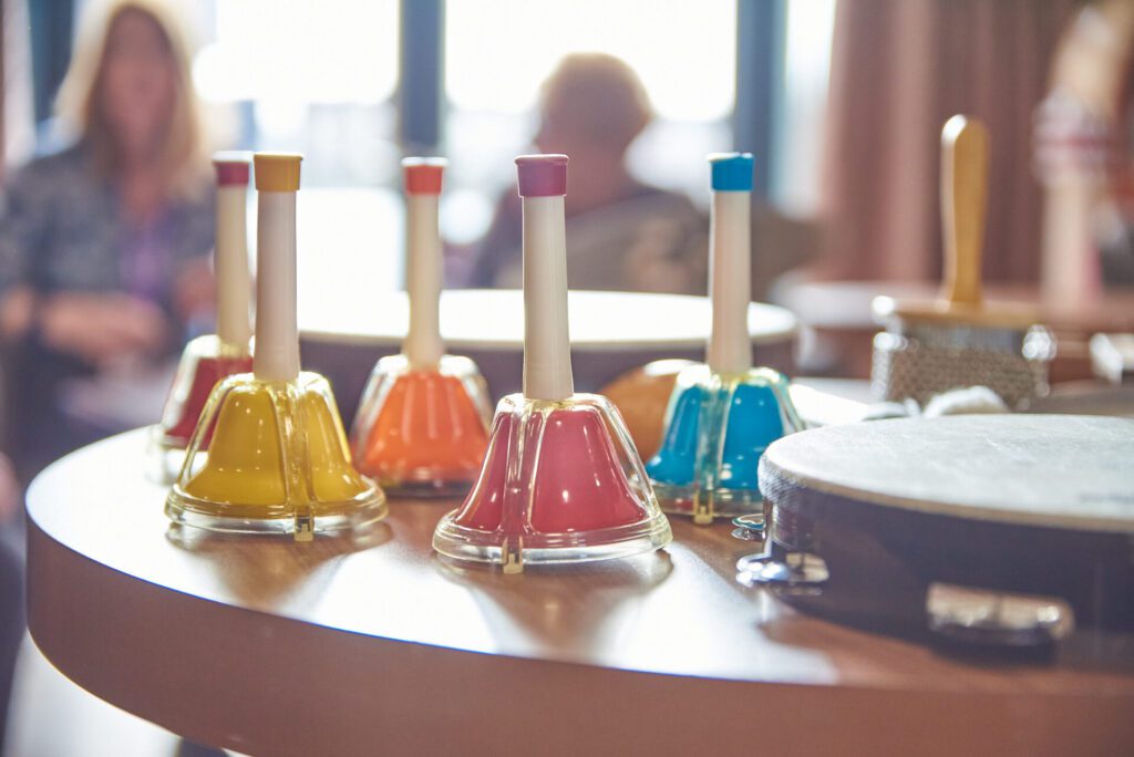A selection of handheld bell instruments in bright red, yellow, orange, pink and blue are on top of a table next to a tambourine. The background of the image is deliberately blurred but there are some people sat down in front of a window.