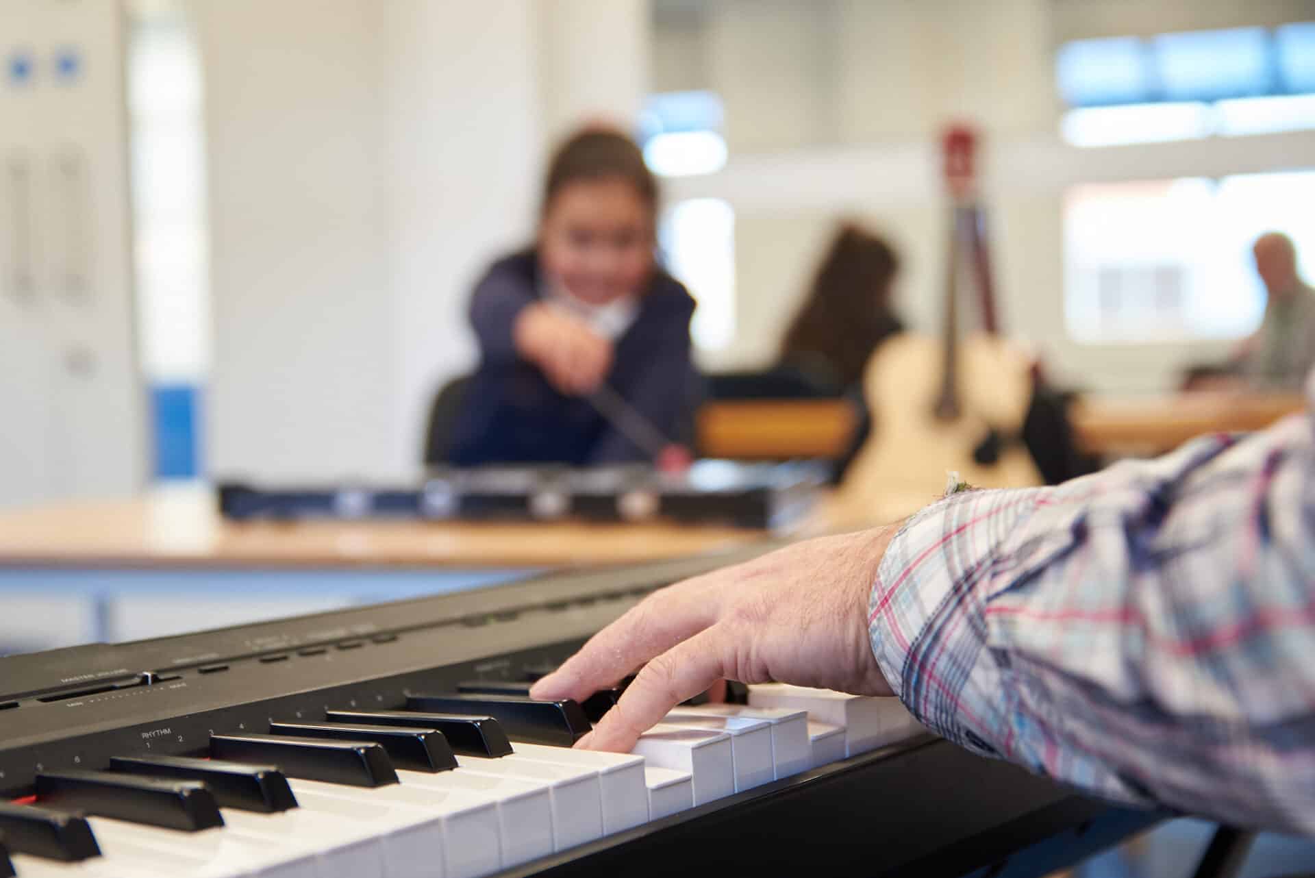 A music therapist plays the electronic keyboard in the foreground, with a child client playing a xylophone in the background.
