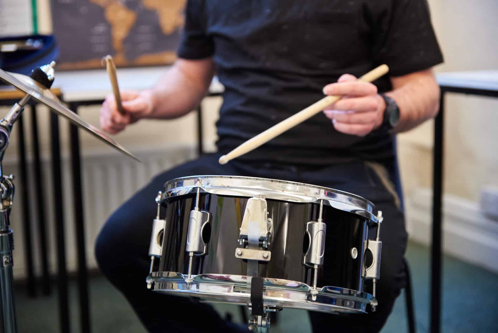 A person sits on a stool playing a snare drum with two drum sticks.