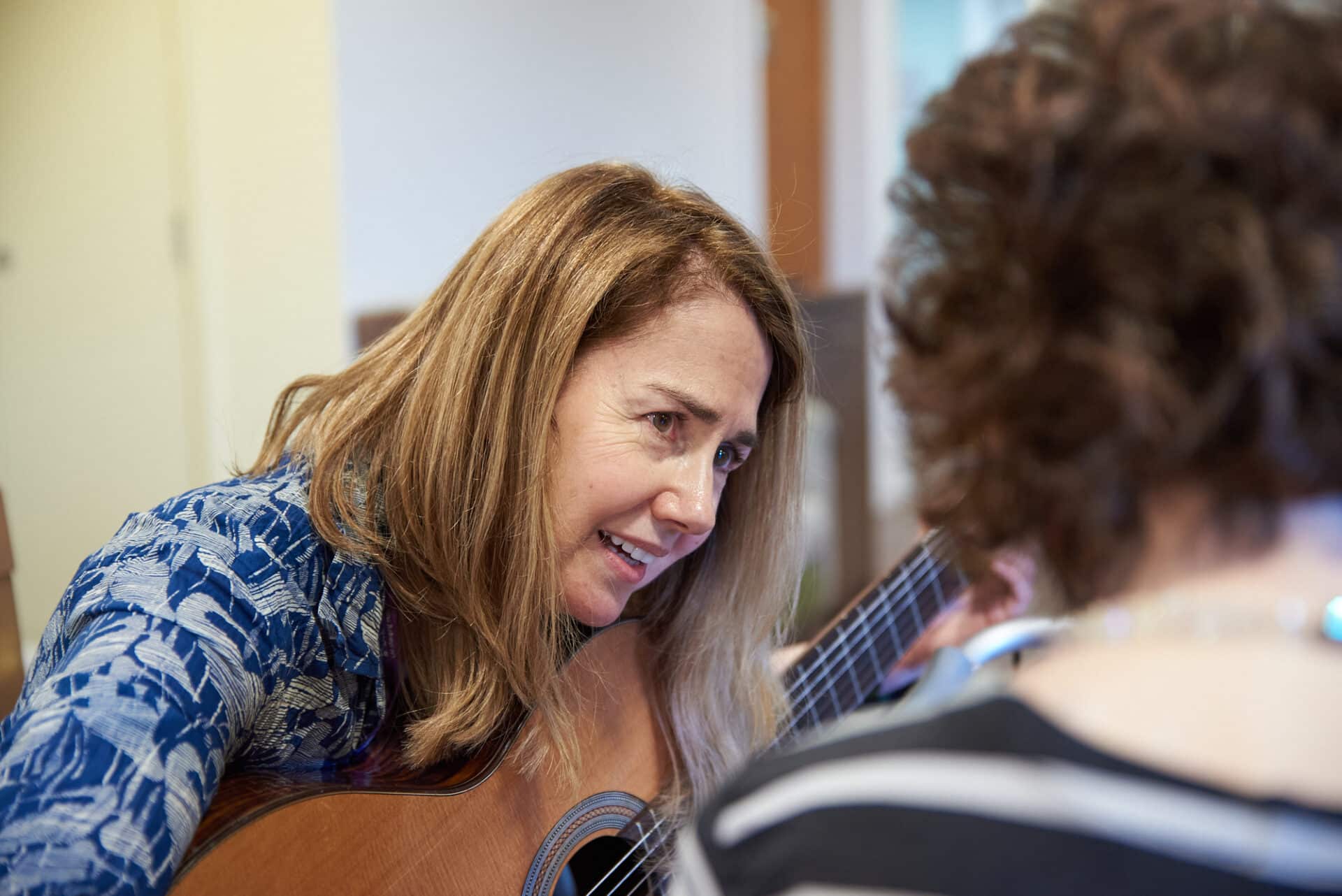 A music therapist leans closer to her client whilst holding an acoustic classical guitar.