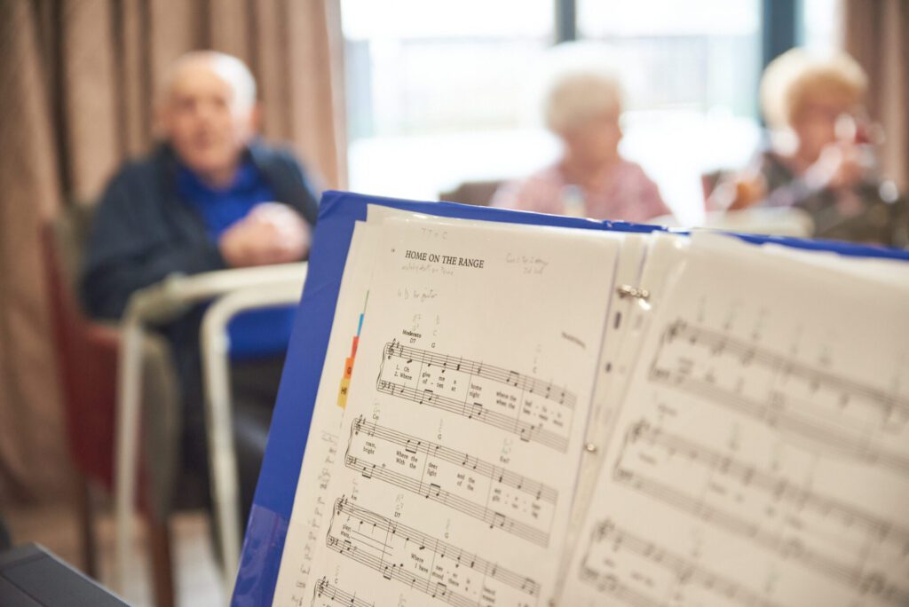 A folder of sheet music is open, with a group of people in the background during a music therapy session.