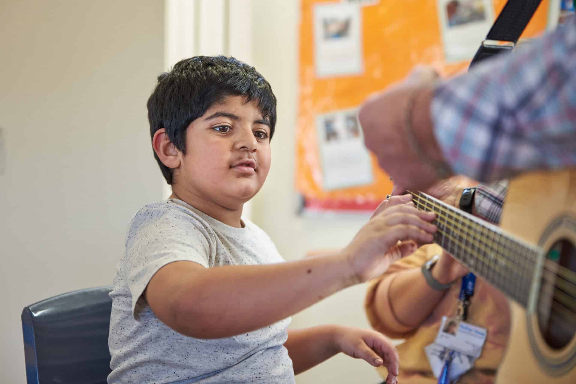 A child client reaches out to touch the strings of the guitar his music therapist is holding.