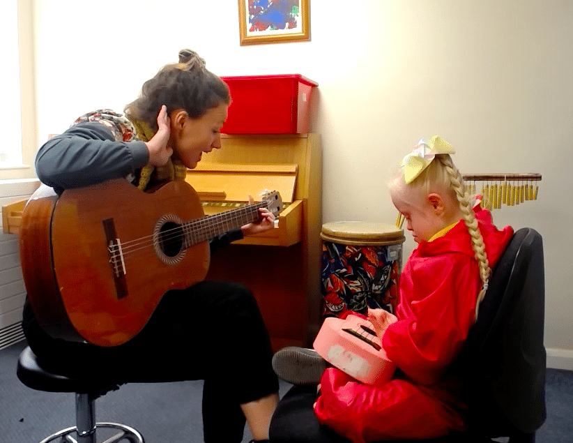 Music therapy takes place at Hadrian school. One of our music therapists plays the guitar, as a young pupil strums a pink ukulele.