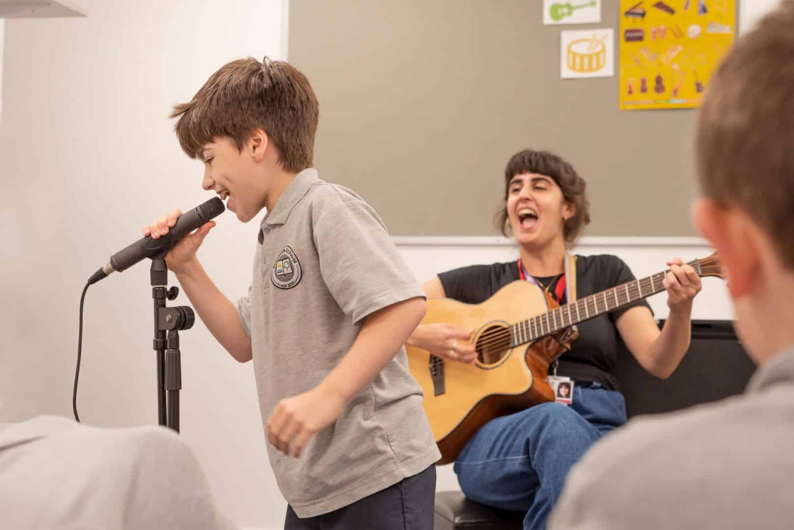 A pupil from the Riverbrae School Choir sings into the microphone, as music therapist Stella plays the guitar.