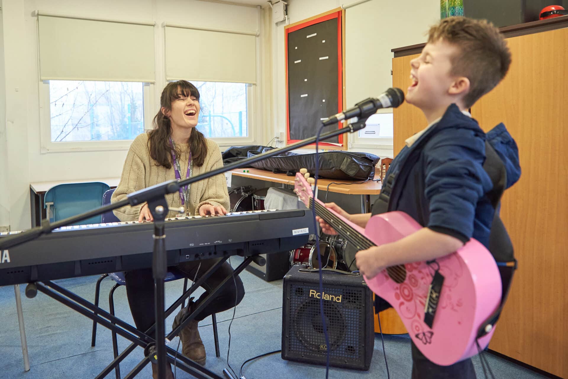 A young woman with long brown hair is playing a keyboard and smiling. In front of her is a school pupil wearing a blue hoodie, holding a pink guitar and singing into a microphone. They look like they are having a lot of fun playing music together.