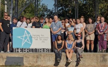 Supporters from The BRIT Trust gather around a sign which says: Improving lives through the power of music and the creative arts.