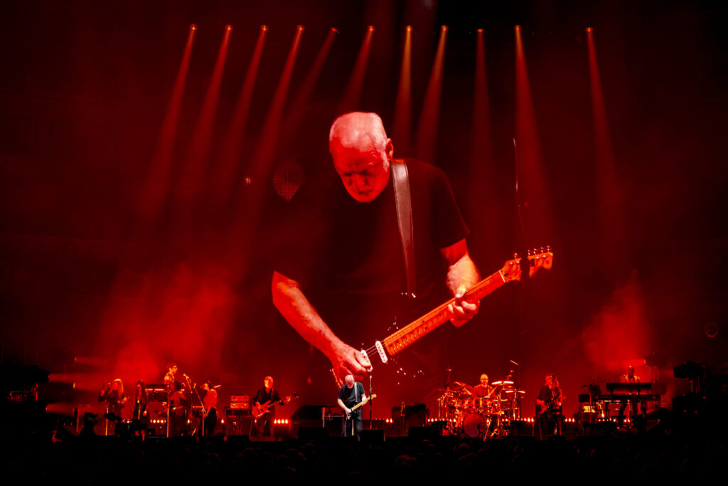 David Gilmour playing guitar on stage against a red background