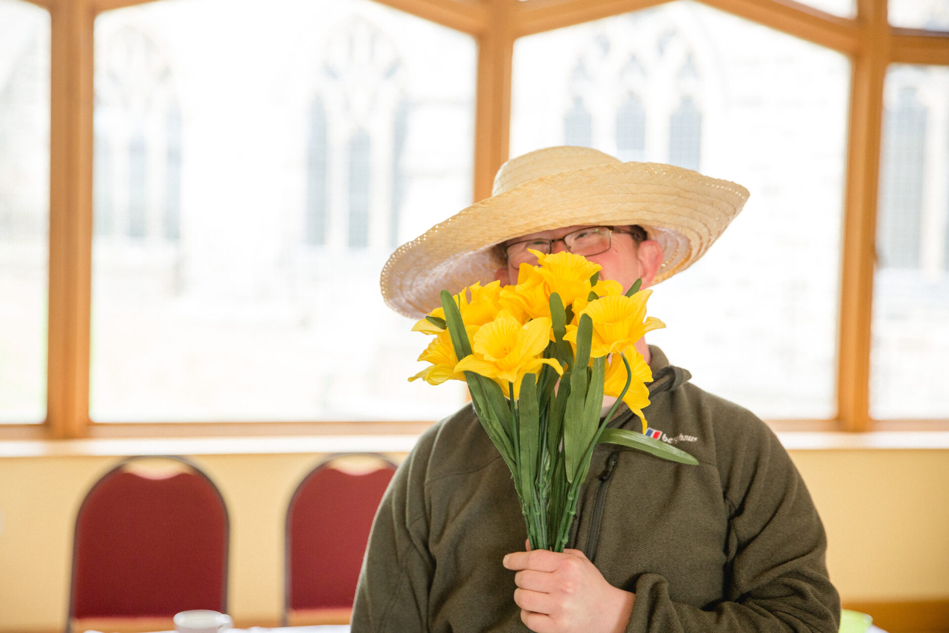 A young man wearing a large brimmed hat is holding a bunch of yellow daffodils in front of his face.