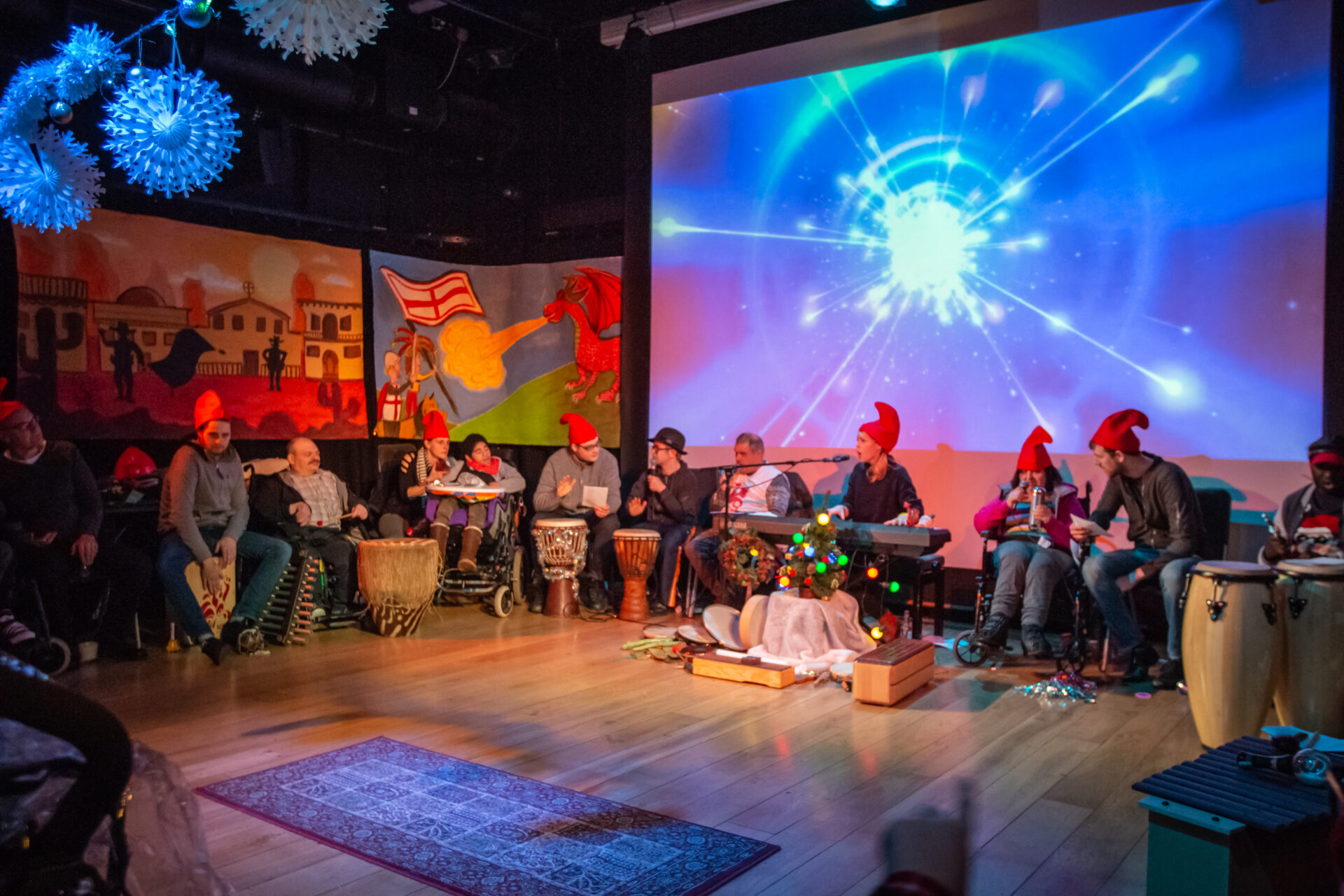 A group session where people are sat in a big circle with instruments, such as big drums, in front of them.