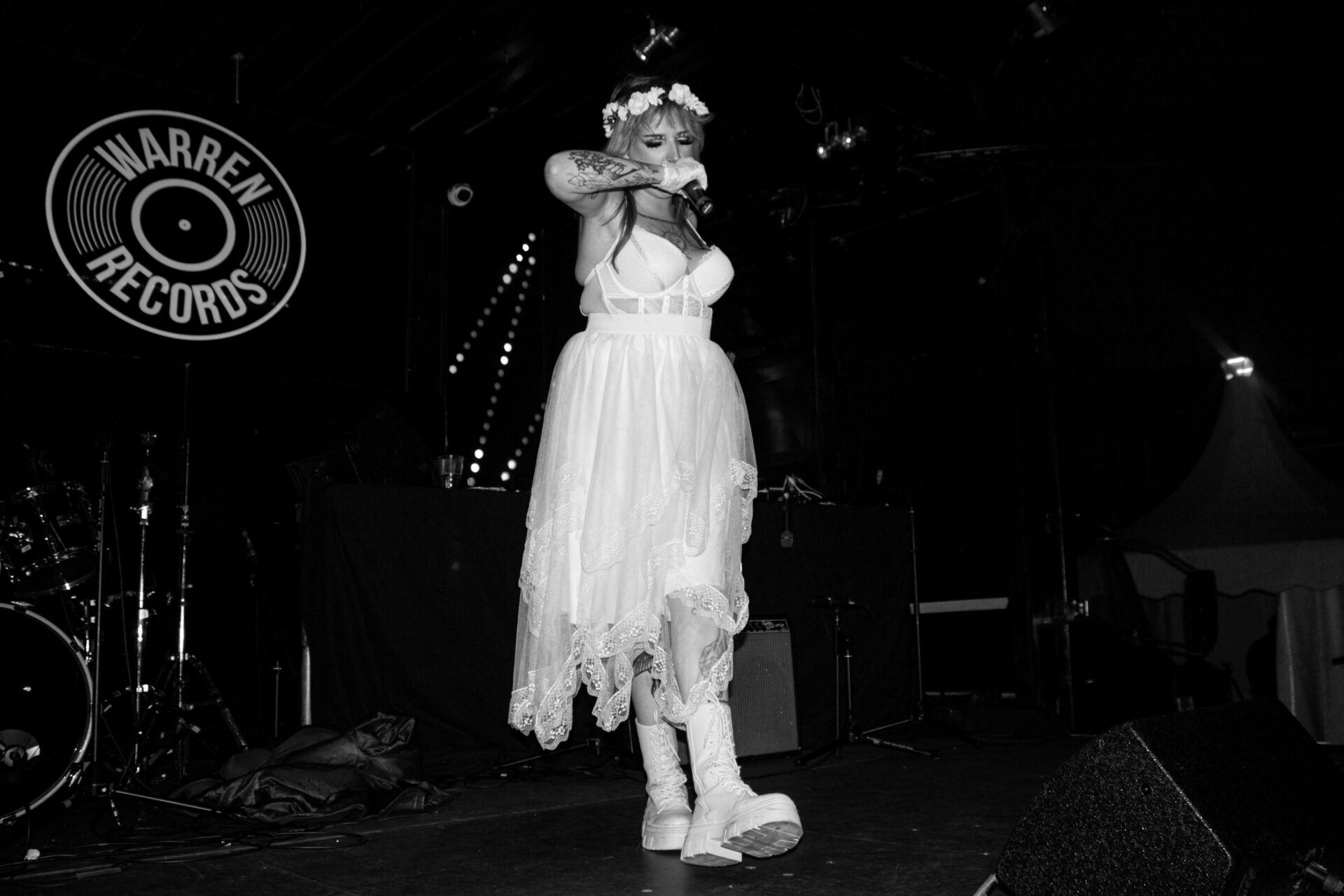 A young woman on the stage in a white dress and chunky white boots is singing into a microphone.