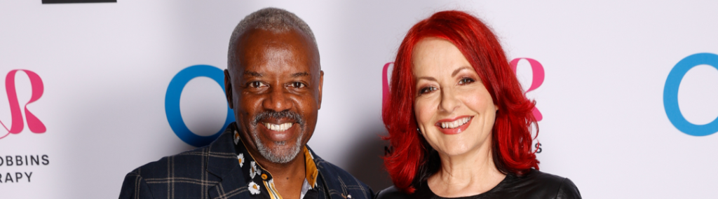 David and Carrie Grant stood, arms around each other, smiling in front of a sponsor board. David is wearing a checked suit, Carrie is wearing a black dress and has red hair.