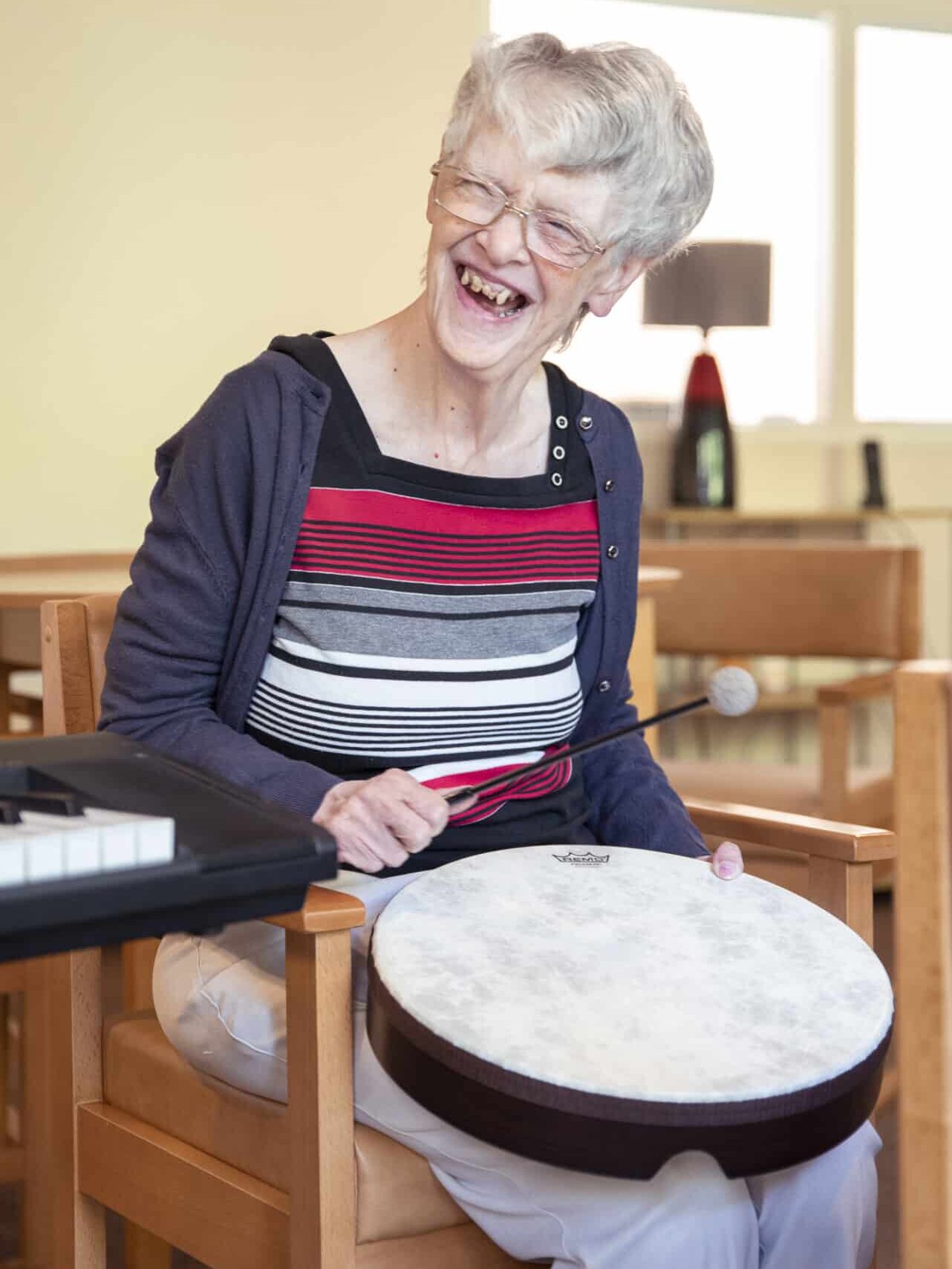 Janet laughs and smiles during a music therapy session, looking to her right as she hits a drum with a drumstick.