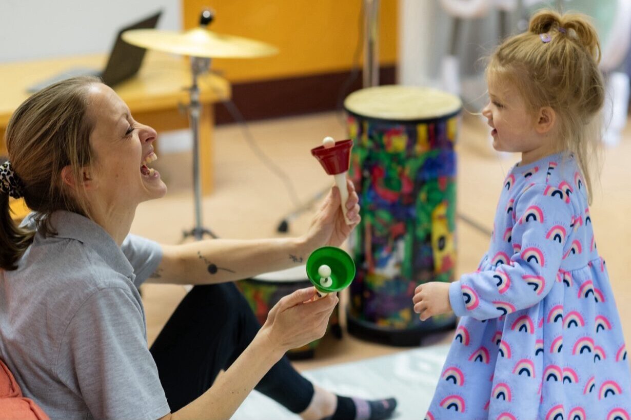 Poppy in a music therapy session, smiling and engaging with a music therapist who is sat on the floor with some bells.