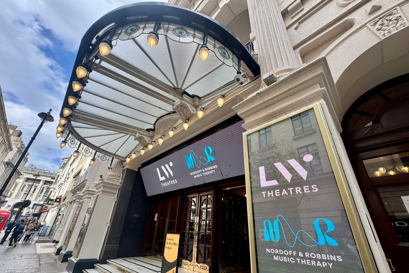 The entrance to historic LW Theatres venue 'The London Palladium', featuring ornate columns, a curved marquee with lights, and digital signage reading "LW Theatres" and "Nordoff and Robbins Music Therapy."